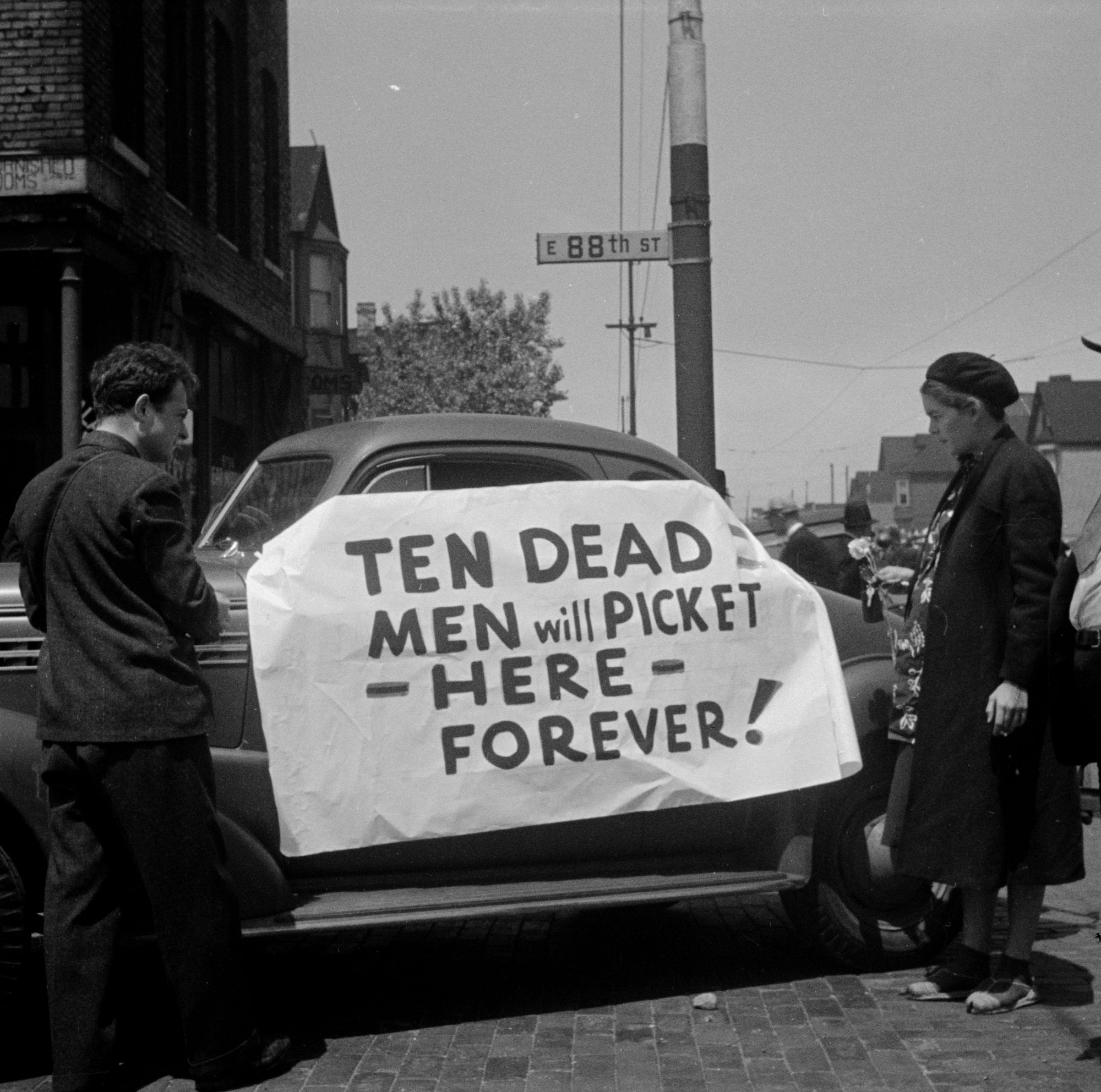 A woman and a man, who is possibly a photographer, looking at a sign on an automobile at the Republic Steel strike in Chicago, Illinois, 1937. One of a set of images showing police officers, politicians, and other people doing activities related to the strike.