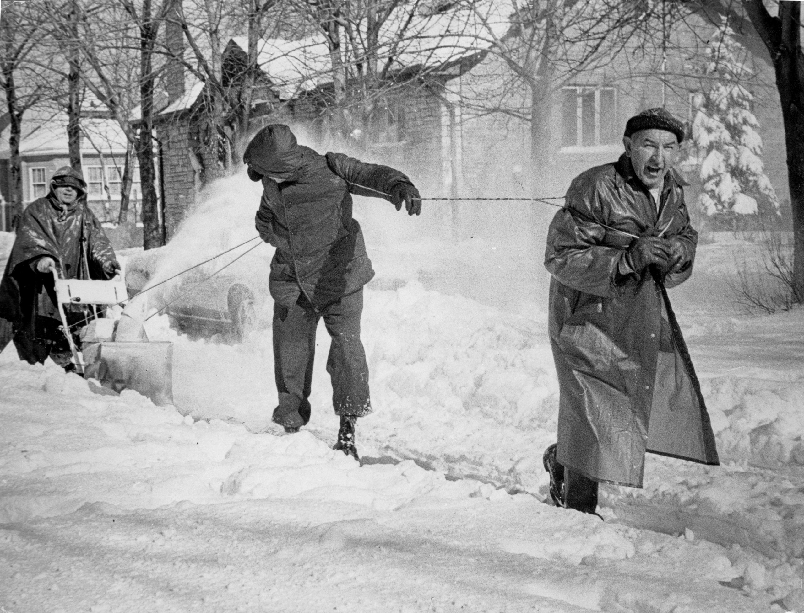One man pushes a snowblower while two men pull with rope to clean the sidewalk after the blizzard of 1967