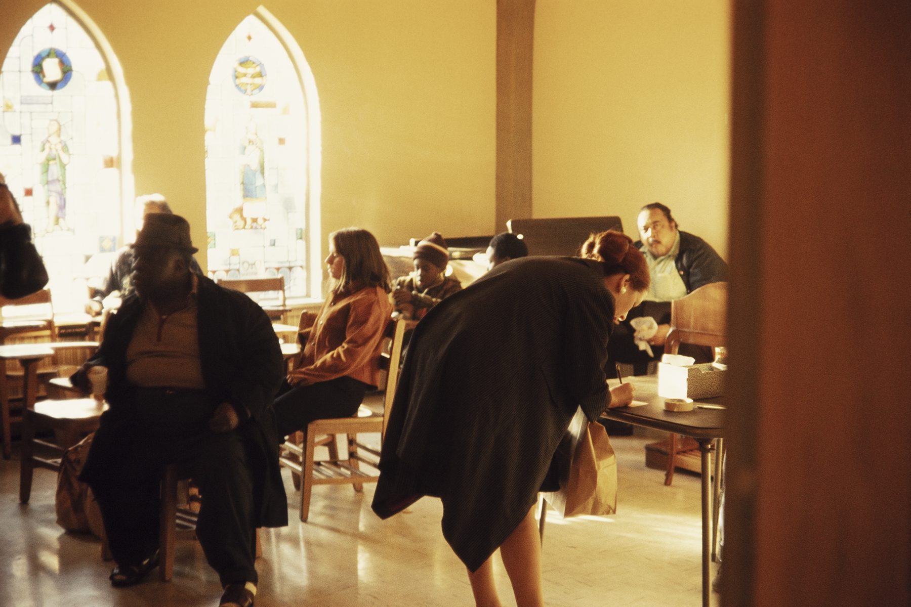 Color photograph of a sun-lit room with 2, stained-glass windows with tapered tops. There are chairs and tables in the room, with 7 seated people. A woman in the foreground bends over with her back to the camera to write something on a piece of paper on a table.