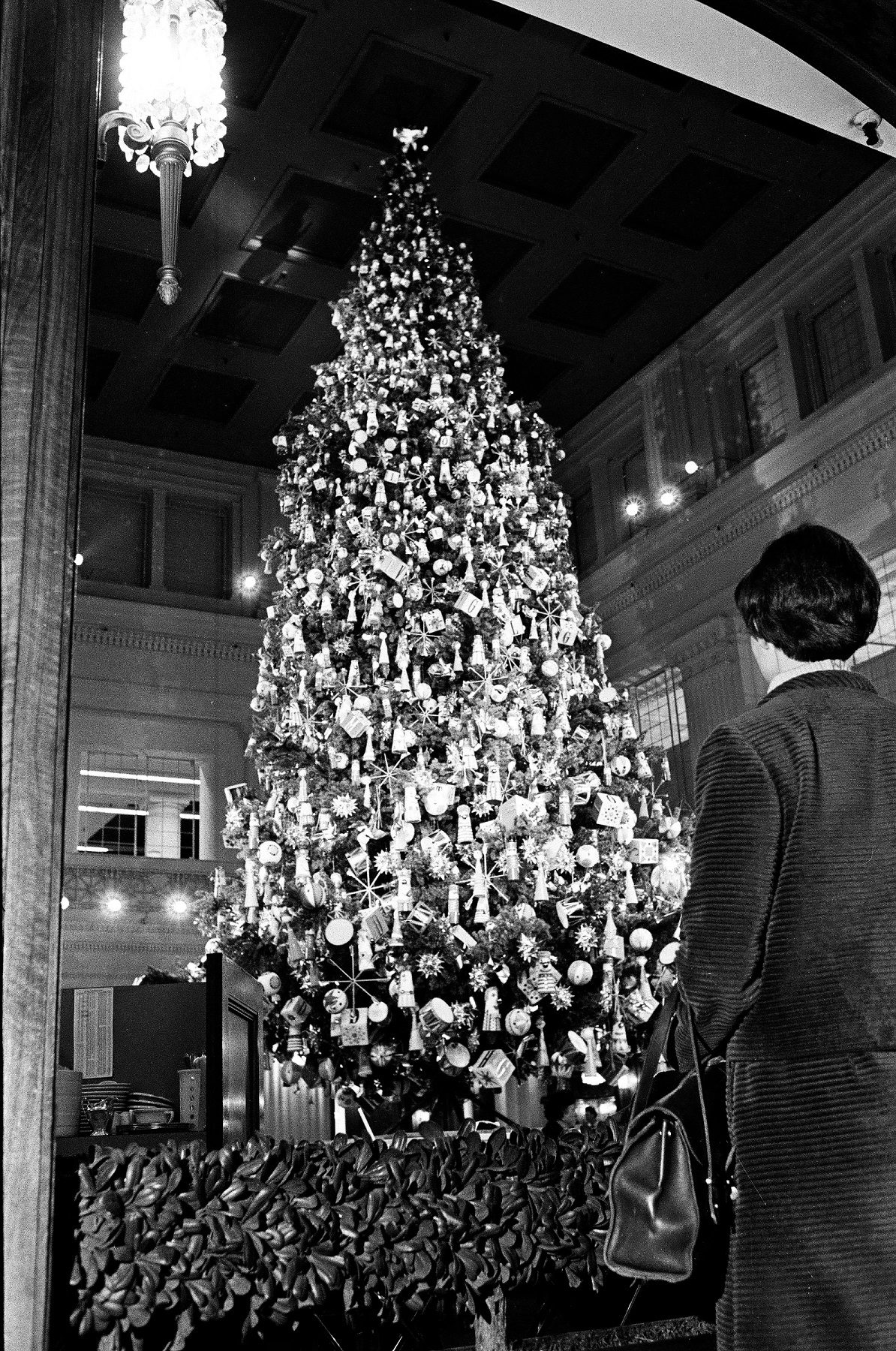 A black & white photograph of a Christmas tree in a tall, large atrium. In the right foreground is the back of a woman gazing at the tree. She has short, cropped hair and is wearing a coat & carrying a handbag. The tree is full of lights & ornaments.