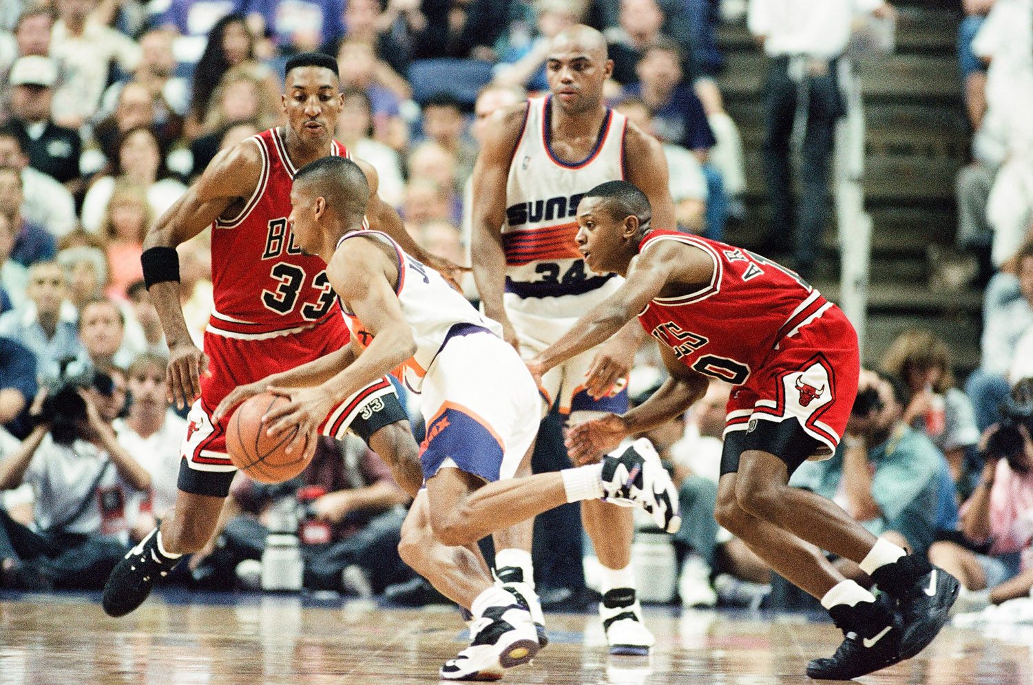 Color photo of Kevin Johnson in a white Suns uniform taking the ball up the court while being guarded by B.J. Armstrong in a red Bulls uniform. Scottie Pippen and Charles Barkley can be seen in the background.