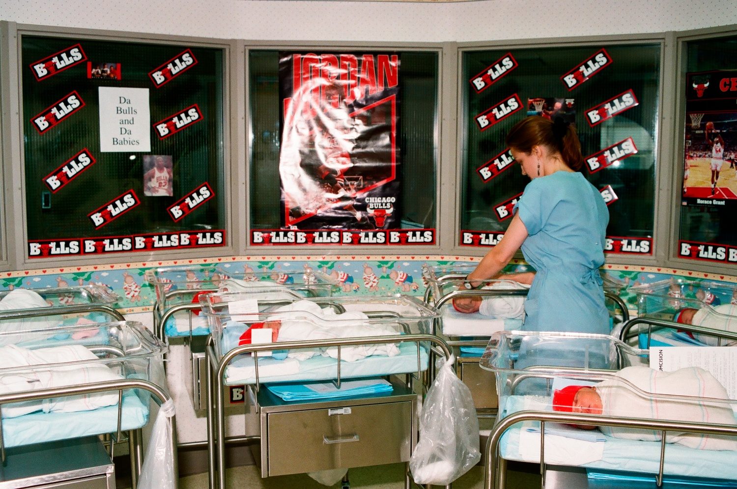 Color photo of a hospital nursery with Bulls posters on the wall and newborns in small cribs wearing red hats as a nurse in blue scrubs tends to them.
