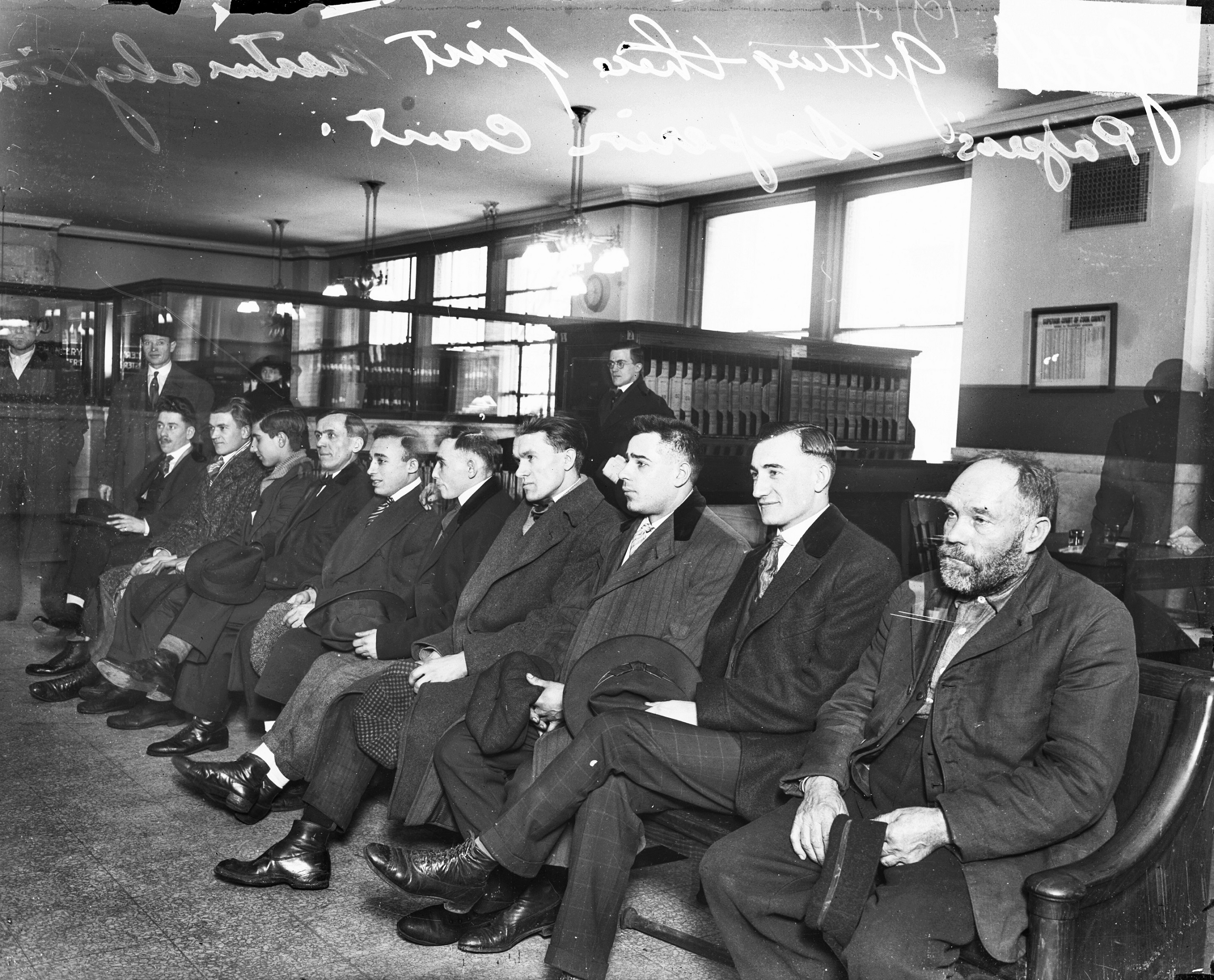 Group portrait of ten immigrant men sitting in a row in the Superior Court on the 4th Floor of the County Building at 118 North Clark Street in the Loop community area of Chicago, Illinois. Text on the image reads getting their first naturalization papers, Superior Court.