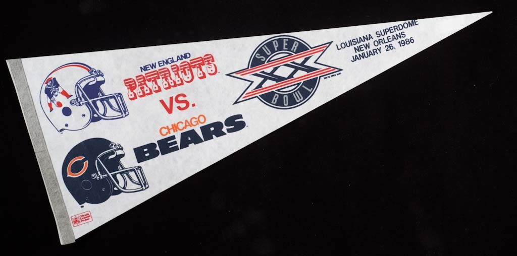 White pennant in triangle shape, with blue, red and orange print. Two helmet m.dngs are depicted and read "New England/Patriots/vs./Chicago/Bears" Superbowl emblem that reads "Super/XX/Bowl/" next to "Louisiana Superdome/New Orleans/January 26, 1986." Pennant was made for Super Bowl XX, won by the Chicago Bears.
