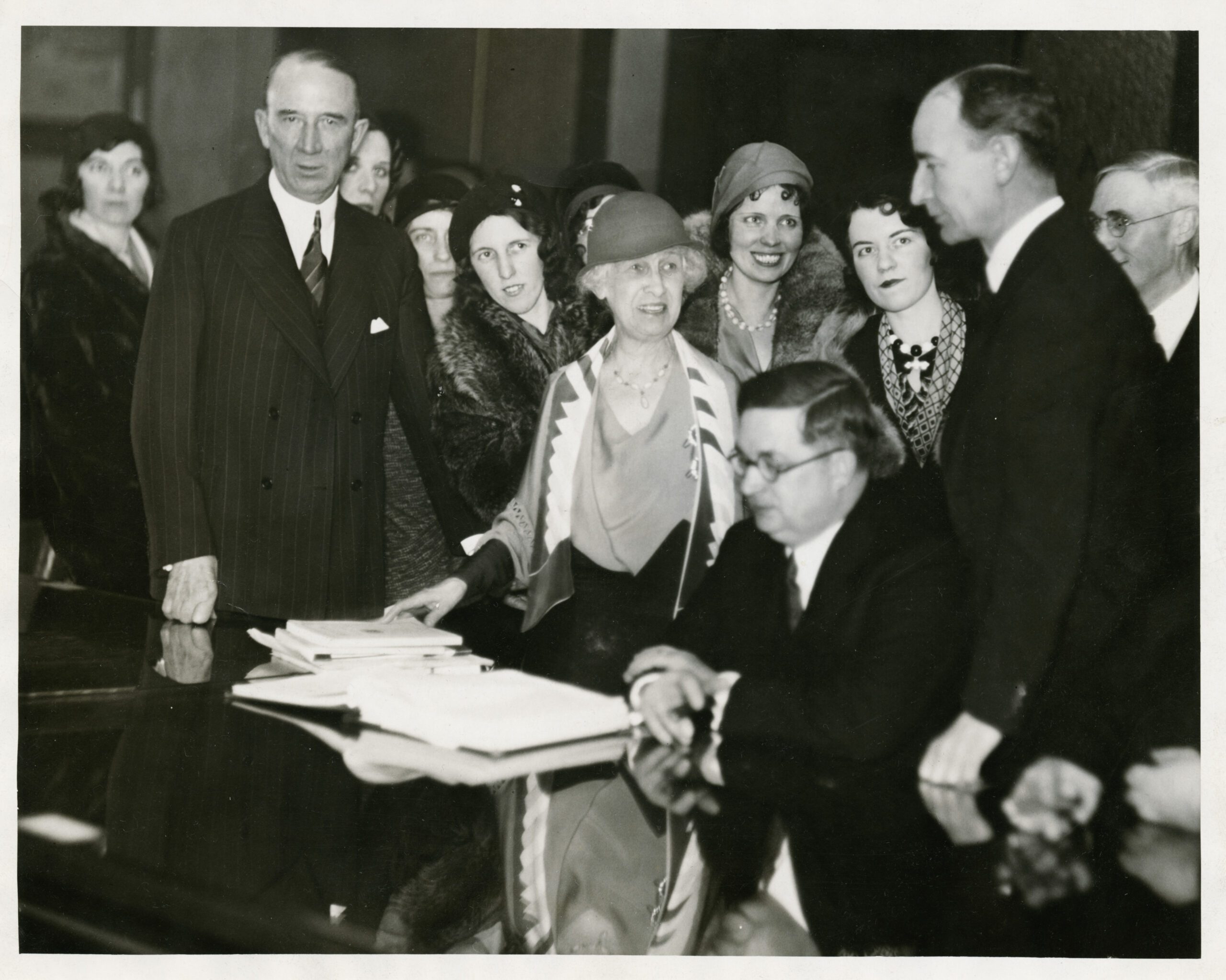 Margaret Haley and several other women stand in a group of men at a desk in 1932