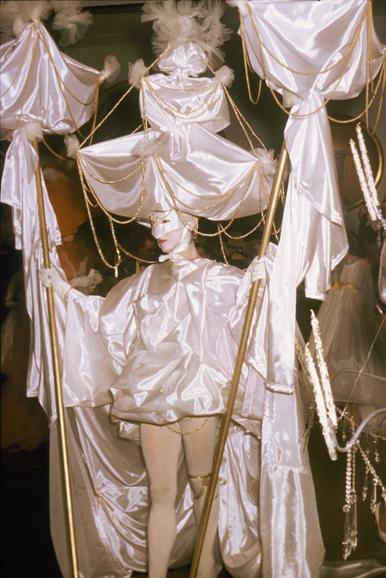 Beaux Arts Ball at the Ambassador West Hotel. Person costumed in white fabric with mask and an elaborate headdress. They are holding two staffs with more swags of white fabric.