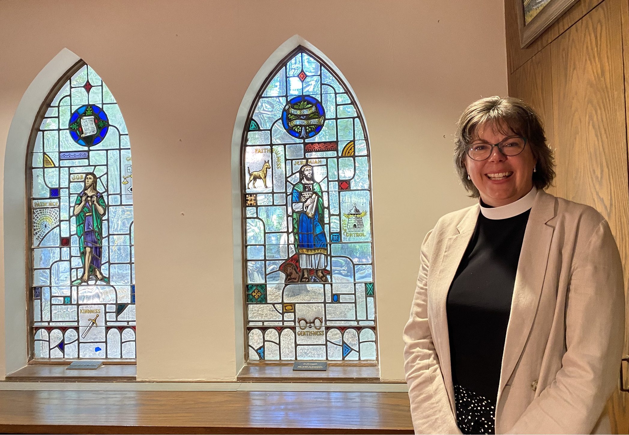 A color photograph of a white woman in a clerical collar and cream blazer standing on the right side of the image with 2 stained-glass windows, one on the left, one in the middle.