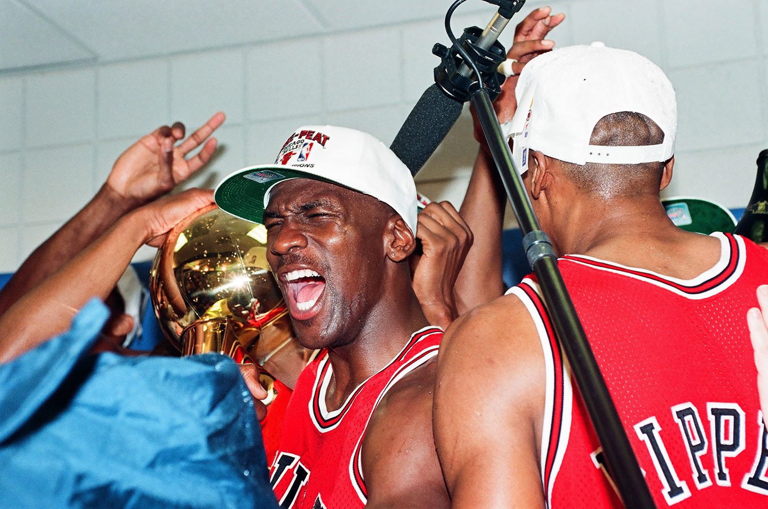 Color photo close up in the locker room of Michael Jordan looking gleefully with the championship trophy in the background and the back of Scottie Pippen visible in the foreground. Jordan is wearing a hat that says 