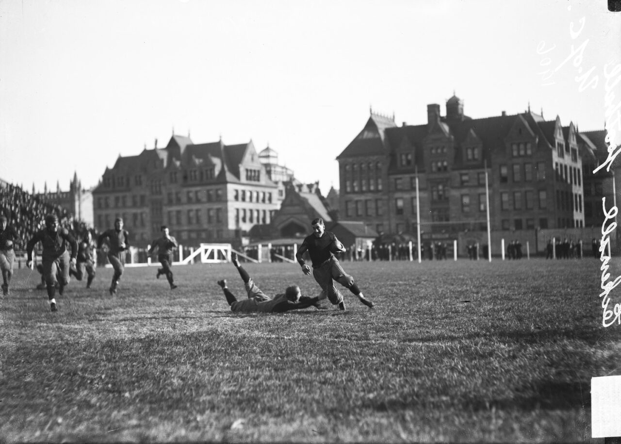 University of Chicago football player Eckersall, cradling a ball in his left arm, running around a downed opponent at Marshall Field