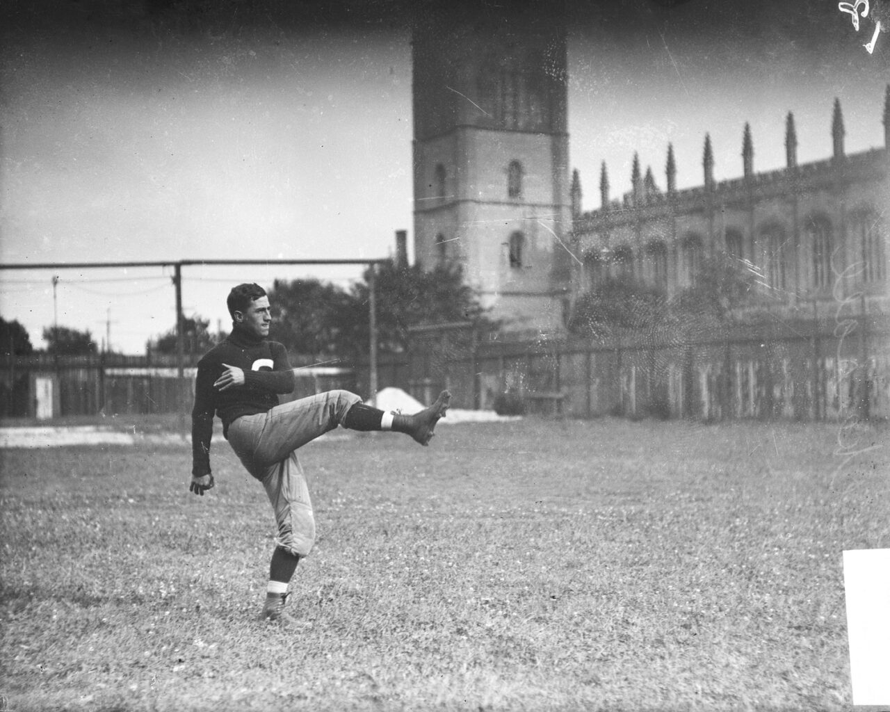 Football player Walter Eckersall holding his leg in the air after kicking a football, standing on the field at Marshall Field